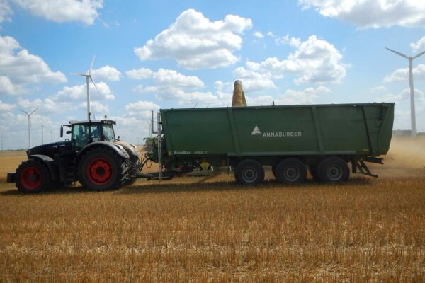 Schwarzer Fendt-Traktor mit grünem Annaburger-Anhänger bei der effizienten Getreideernte auf einem Stoppelfeld, im Hintergrund Windkraftanlagen. Beispiel für moderne, technikgestützte Maßnahmen für Biodiversität und Ressourcenschutz in der Agrarlandschaft.